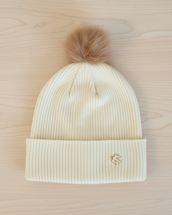 Beige knit beanie with a fur pom-pom on a wooden surface
