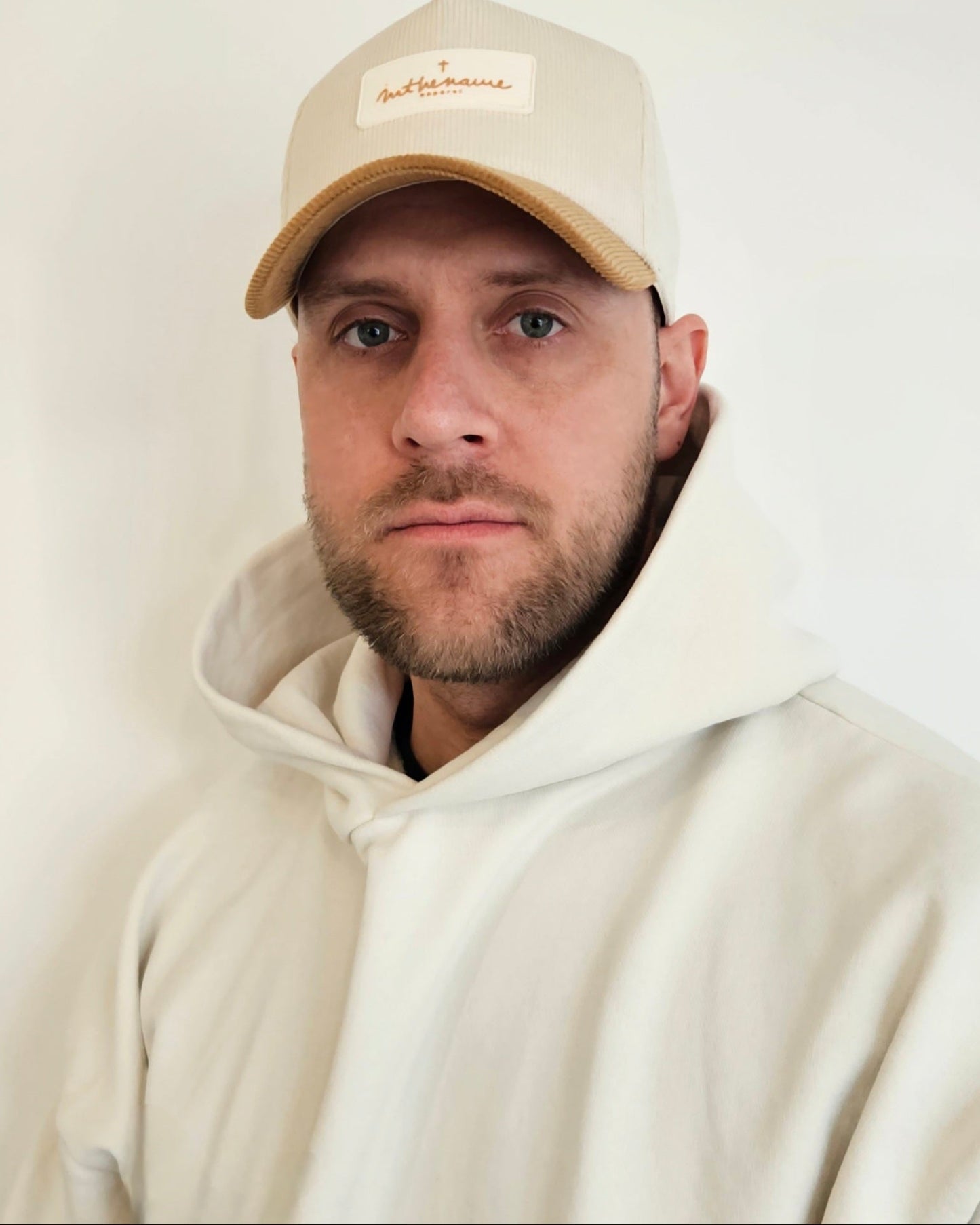 Man wearing a beige cap and white hoodie against a plain background