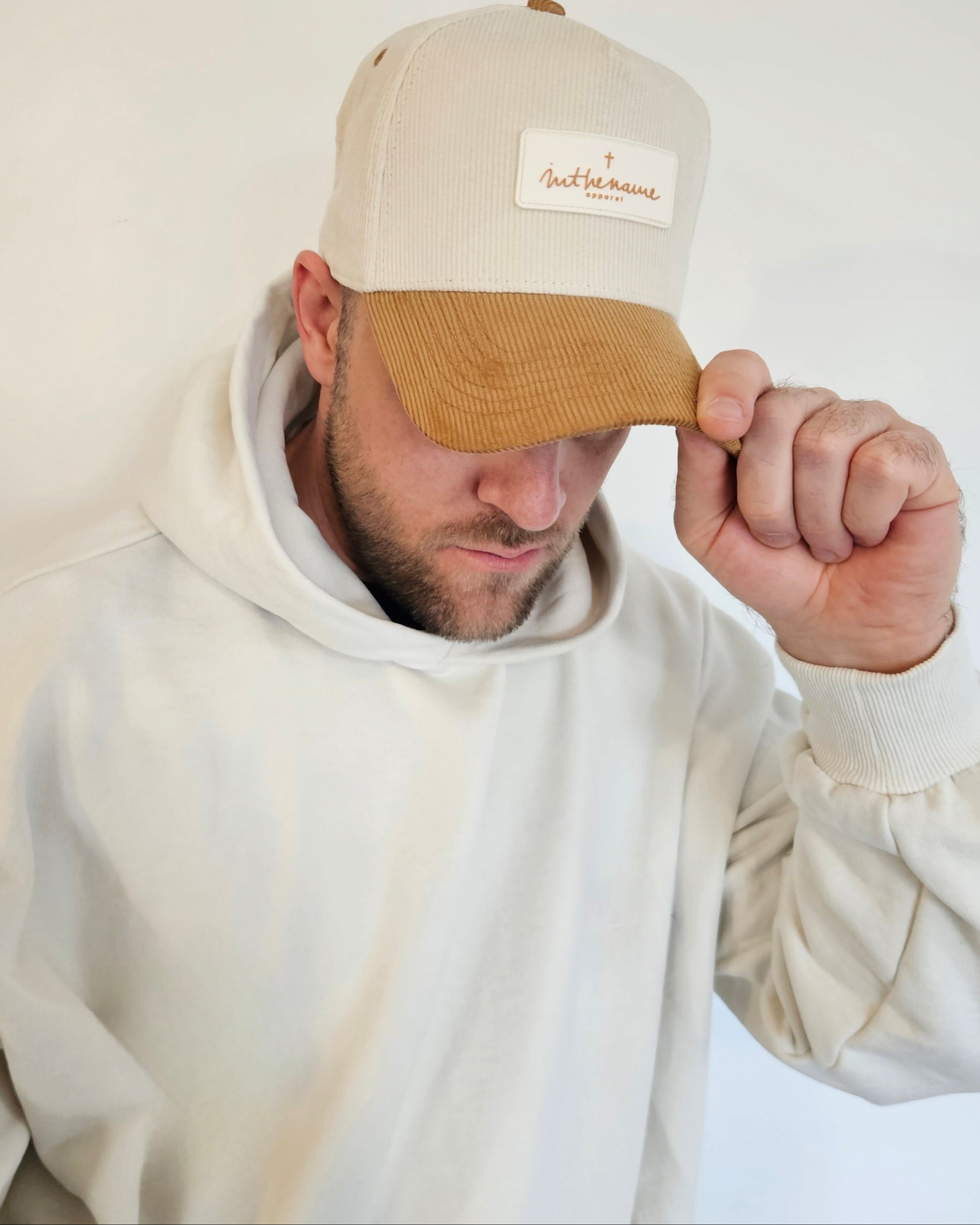 Man wearing a beige cap with a logo and a white hoodie on a light background