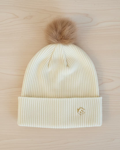 Beige knit beanie with a fur pom-pom on a wooden surface